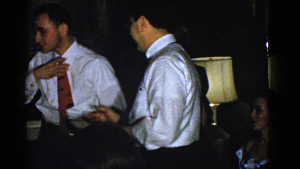 Two men in shirts and ties face each other in the dim party room, with a shaded lamp and seated guest behind them.
