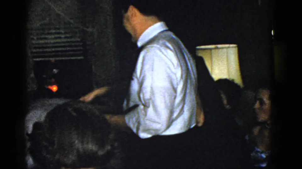 A man in a white shirt stands with his back partly turned as seated guests watch from a dim Boston house-party interior.