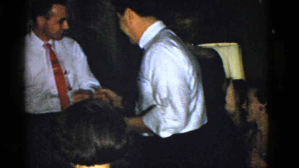 In 1975 Boston home-movie footage, two men in white shirts and ties stand close together at a formal indoor gathering.