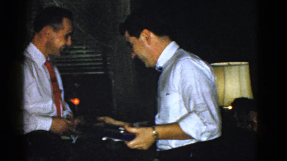 Two men in white shirts and ties appear to exchange a small item during a 1975 Boston indoor gathering.