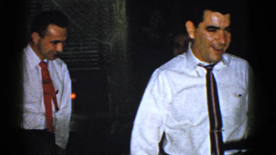 A smiling man in a white dress shirt and dark tie faces the camera during the Boston party scene.