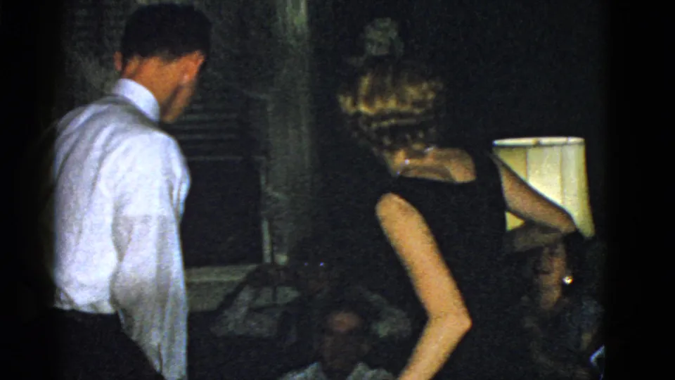 1975 Boston 8mm frame showing adults in formal party clothing inside a dim private room with warm lamp light.