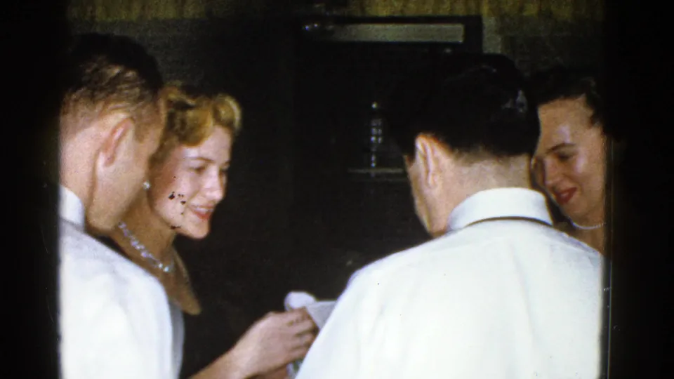 1975 Boston home-movie party frame showing adults in close conversation, with a smiling woman holding a cup.