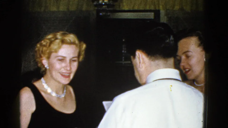 1975 Boston 8mm frame of adults conversing at a dim indoor party, with a woman in a black dress and pearls smiling.
