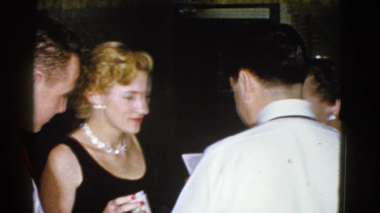 A 1975 Boston indoor gathering shows a woman in a black dress and pearls looking down while several adults stand nearby.