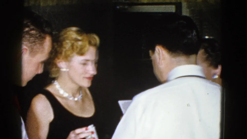 A 1975 Boston indoor gathering shows a woman in a black dress and pearls looking down while several adults stand nearby.