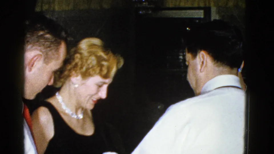 The 1975 Boston home-movie frame shows a woman in black and pearls smiling downward among adults in a shadowed interior.