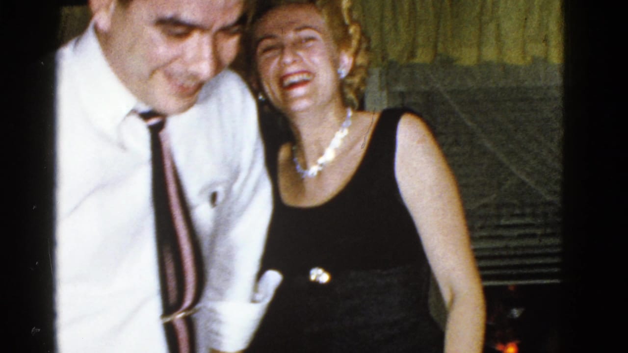 In a 1975 Boston home-movie frame, a man in a white shirt leans toward a laughing woman in a black dress and pearl necklace.
