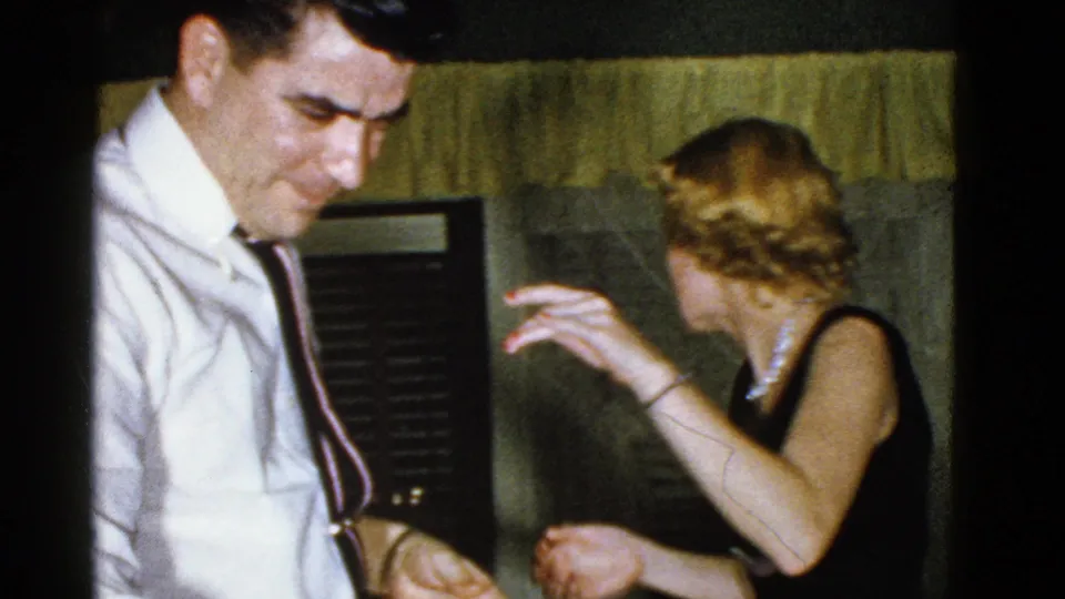 The man looks down as the woman turns and lifts a hand near her hair during the 1975 Boston home gathering.
