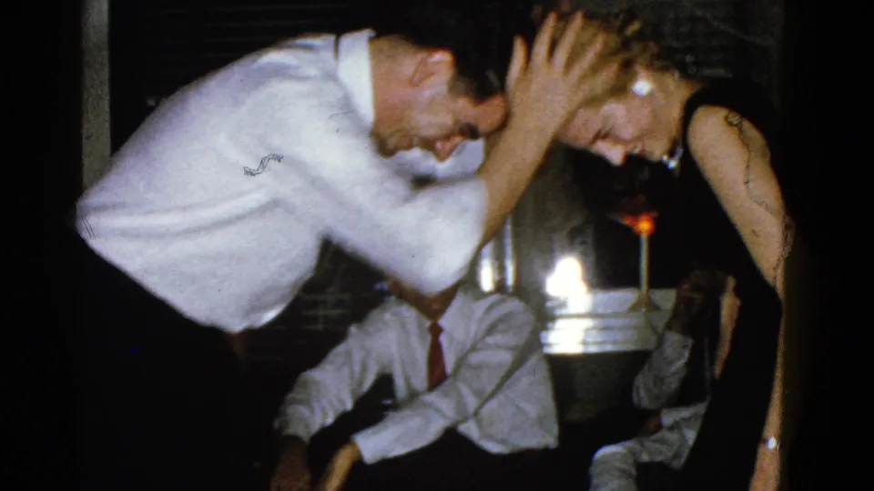 A man in a white shirt leans close to a woman in a black dress during the candle game, with seated adults visible in the dim room.