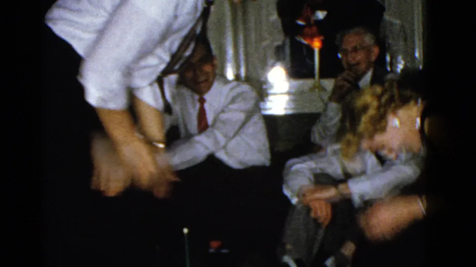 Adults in dress clothes crowd around the tabletop game in a 1975 Boston home-movie party scene, with a candle and small upright piece visible.