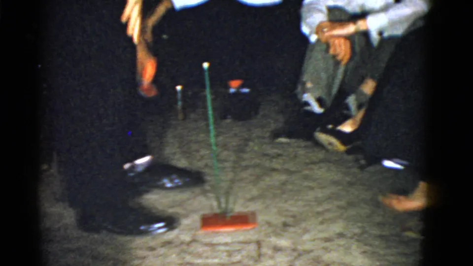 The camera frames the lower half of party guests around a candle and upright game piece, emphasizing the informal play in a private interior.