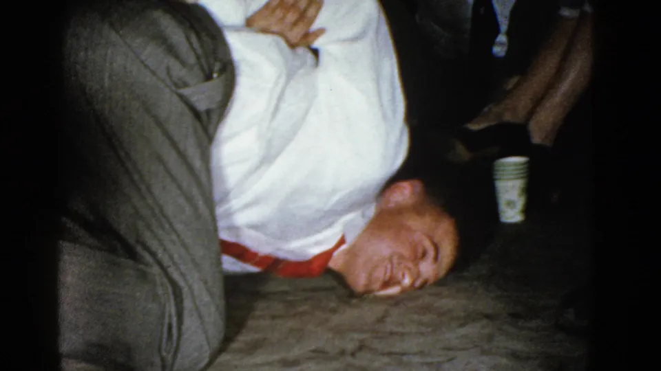 In a 1975 Boston home-movie frame, a man in a white shirt and red tie is held down near the floor during playful wrestling at an indoor gathering.
