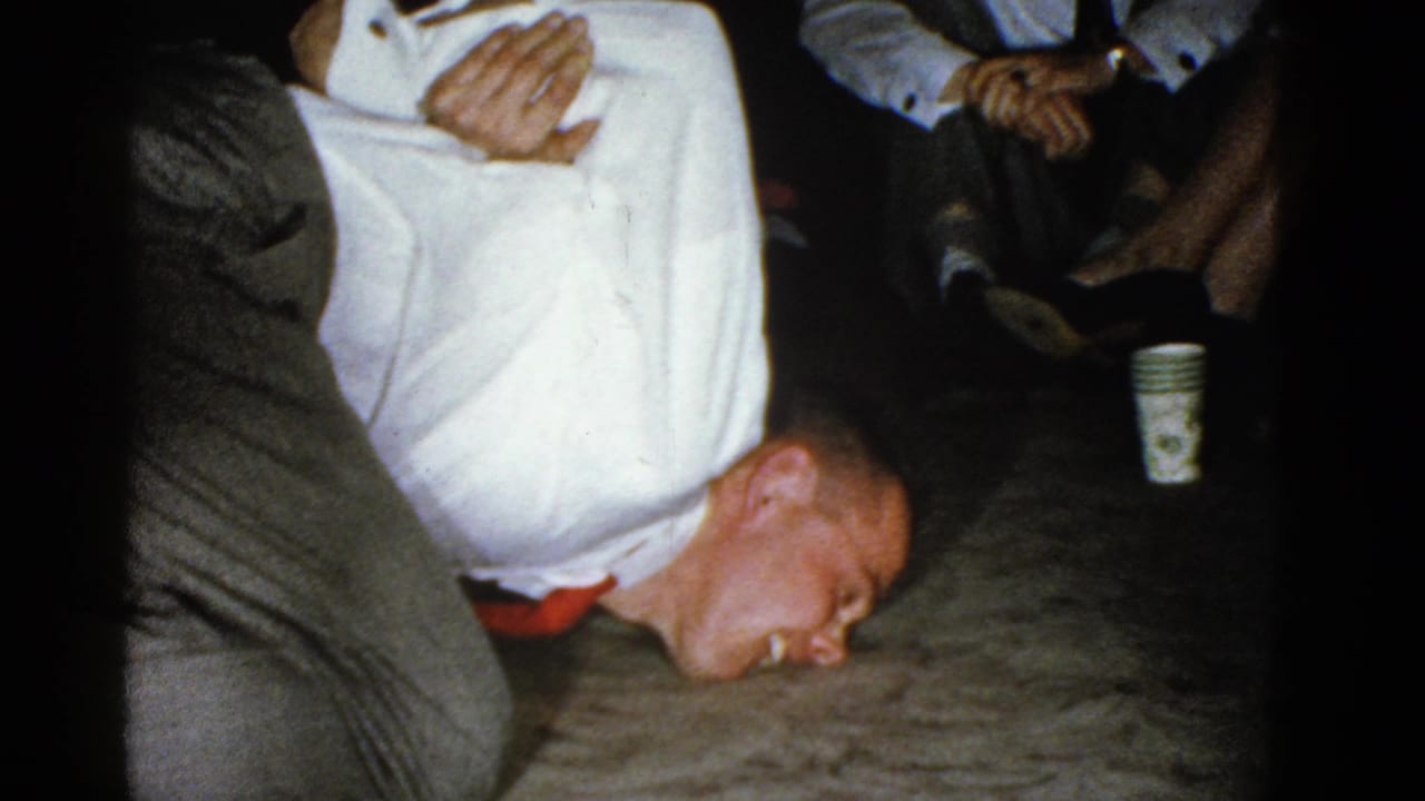 Man lies face down near the floor with another person holding him as seated adults watch.