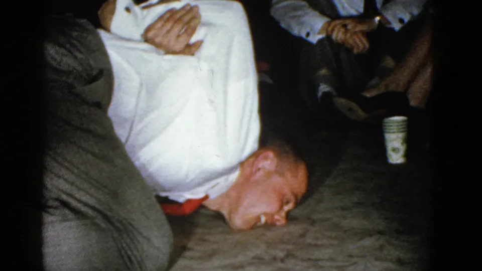 A man in a white shirt and red tie lies face down near the floor while another adult holds him during a casual 1975 Boston indoor gathering.