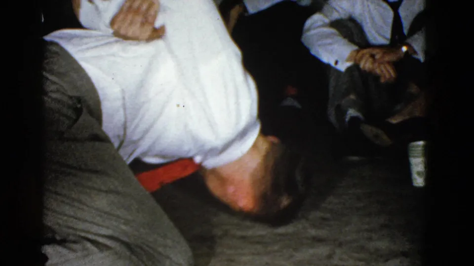 The handheld 8mm frame shows a close, partly cropped view of the wrestling moment, with the man’s white shirt and red tie visible near the floor and seated adults in the background.