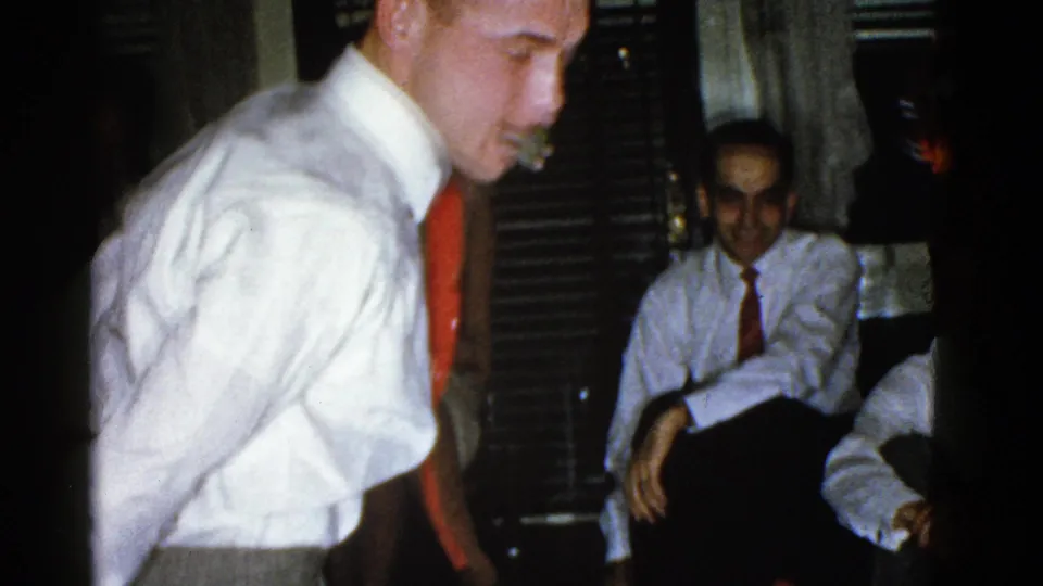 A man in a white shirt and red tie rises from the floor as seated men watch in a dim private interior, consistent with the 1975 Boston home-movie gathering.