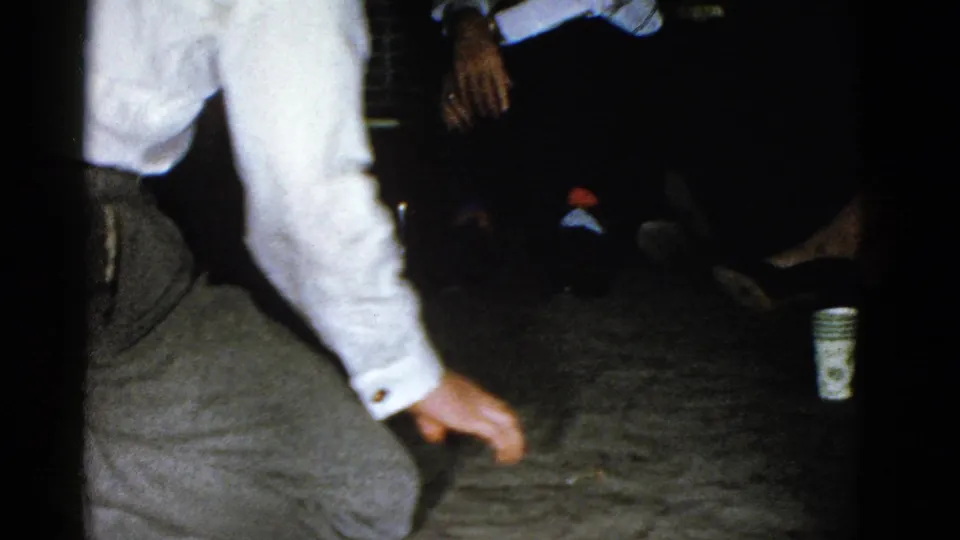 A dim 1975 Boston party interior shows seated guests, a paper cup on the floor, and the partial figure of a man continuing the playful action.