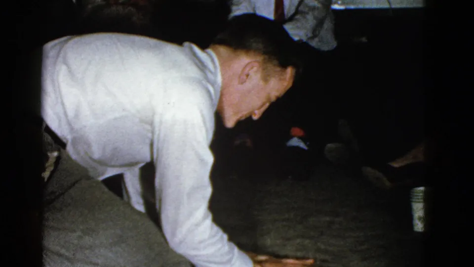 In the final 1975 Boston party frame, the man leans low and reaches toward the floor as seated guests remain gathered around the room.