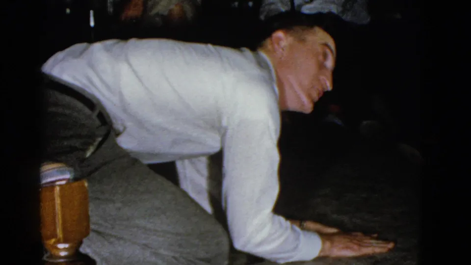 A close 1975 Boston home-movie frame shows the kneeling man beside a wooden chair leg, leaning toward the floor in a dim room.