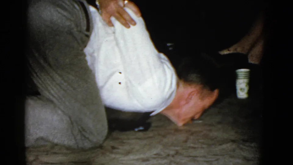 The man leans close to the carpet in a dim private interior, with another person steadying him during the party game.
