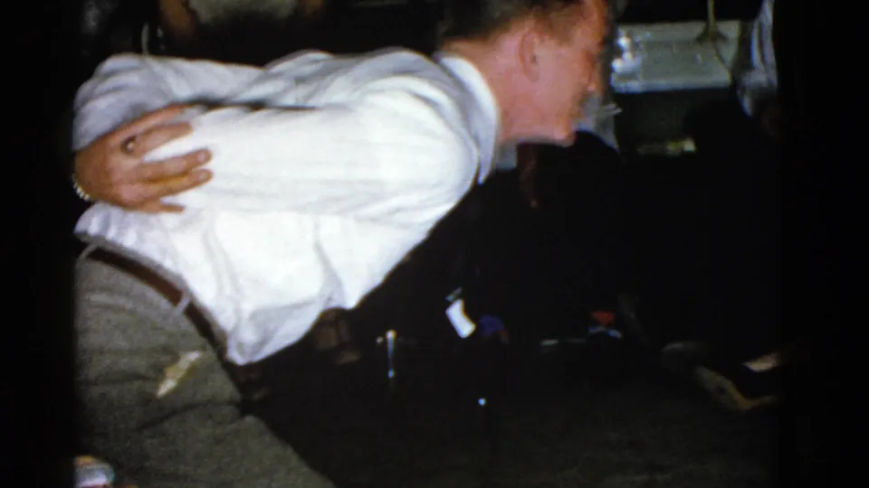 The participant bends sideways in the private indoor gathering, with seated adults partly visible in the dark background.