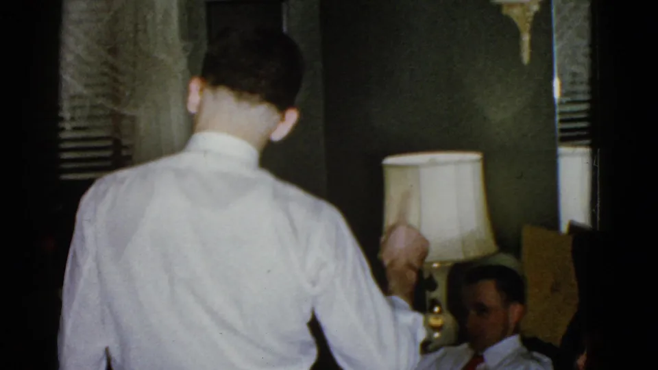 In a dim 1975 Boston interior, a man in a white dress shirt gestures toward a seated man near a table lamp and blinds.