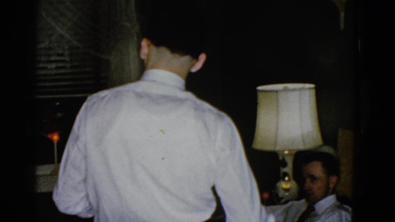 A man in a white shirt faces away from the camera in a dim Boston room, with a seated man partly visible near a lit table lamp.