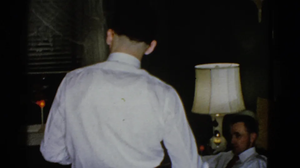 A man in a white shirt faces away from the camera in a dim Boston room, with a seated man partly visible near a lit table lamp.