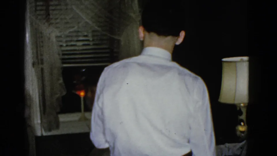 The handheld 8mm frame shows the back of a man in a white shirt near window blinds, curtains, a lamp, and a small candlelit point.