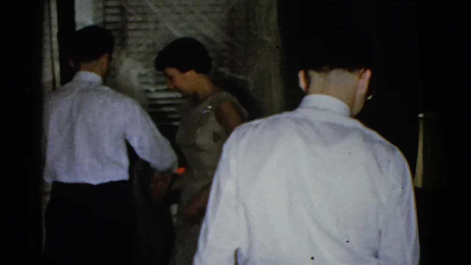 In the final 1975 Boston frame, two men in white shirts and a woman in a dress share a dim private interior near curtains and a lamp.