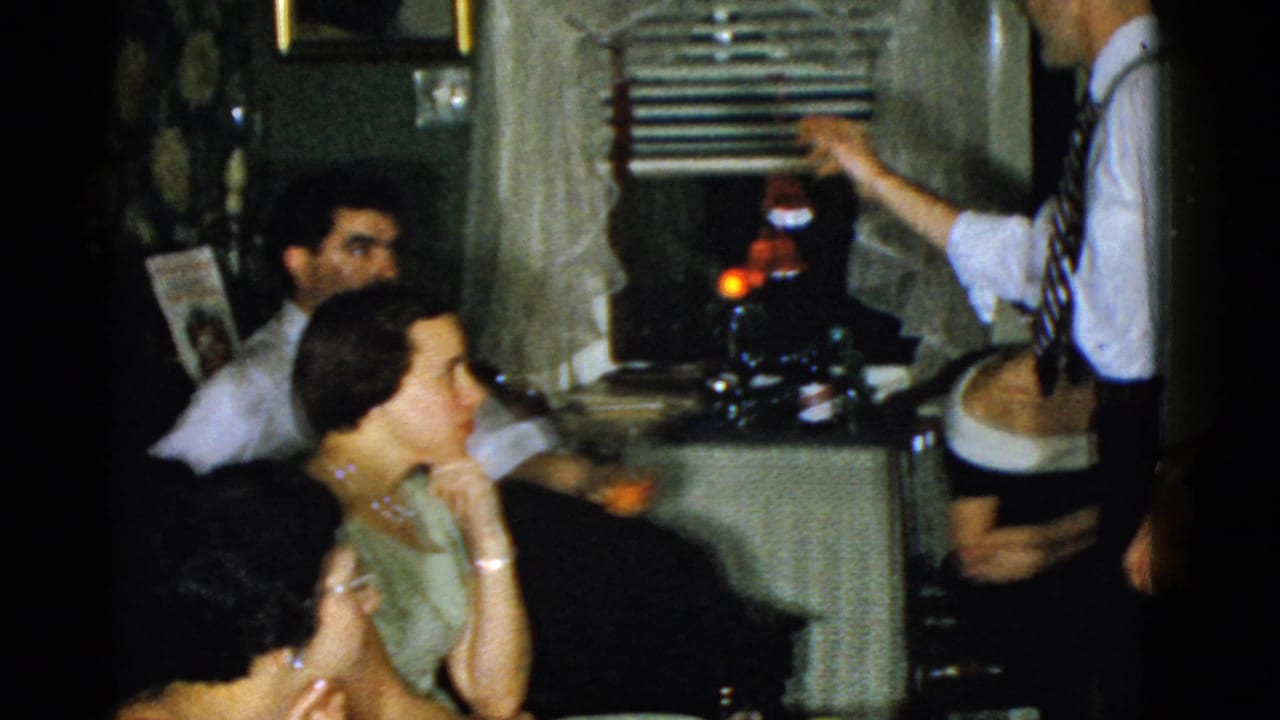 A man in glasses stands near seated adults by window blinds and a side table.