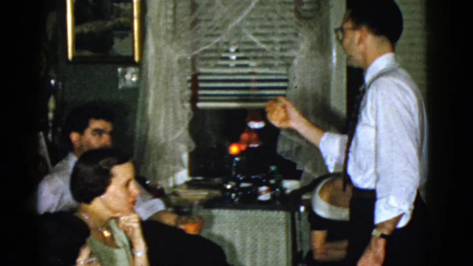 1975 Boston home-movie frame of a man gesturing as adults sit closely around a couch in a private living room.