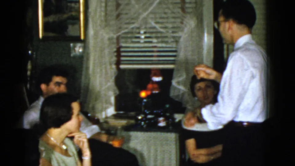 1975 Boston home interior showing a standing man addressing seated adults in a dim living room with curtains and blinds.