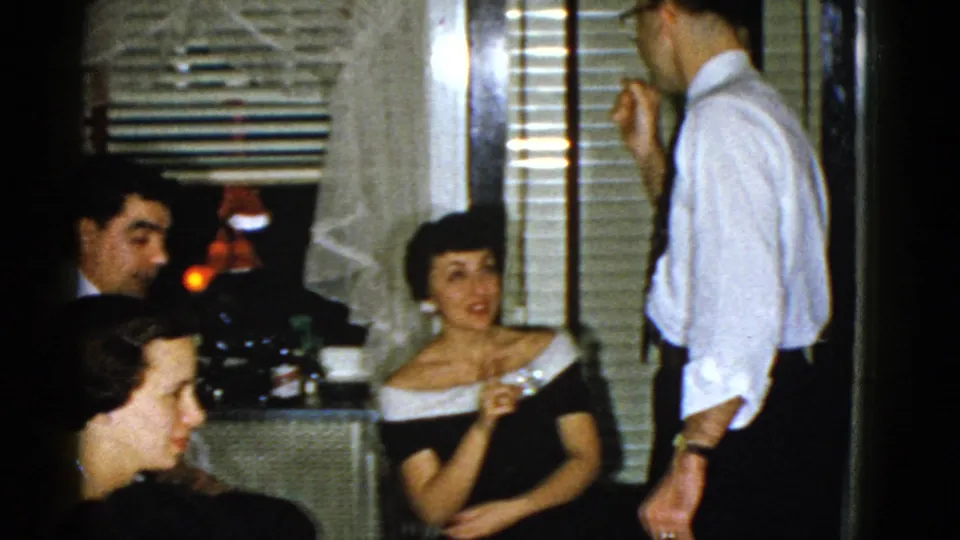 1975 Boston home-movie frame showing adults in a dim private room as a man in a white shirt talks near seated party guests.