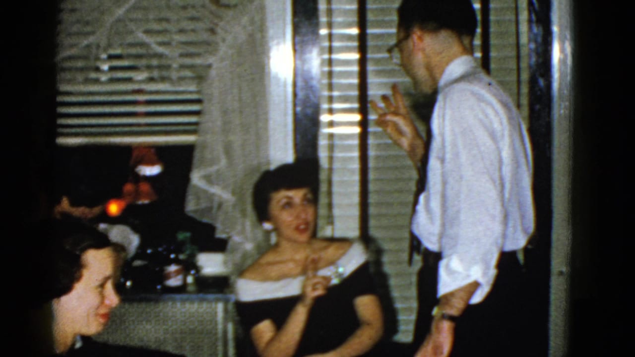 A man in a white shirt gestures while talking to a seated woman at a party.