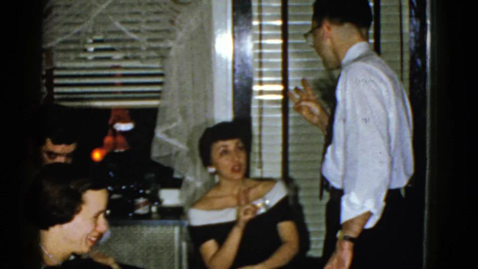 1975 Boston home-movie frame showing seated adults, a woman in formal dress, and a standing man talking in a private interior.
