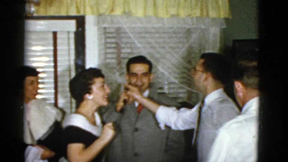 1975 Boston indoor party frame with several adults clustered near curtained windows, smiling and gesturing in conversation.