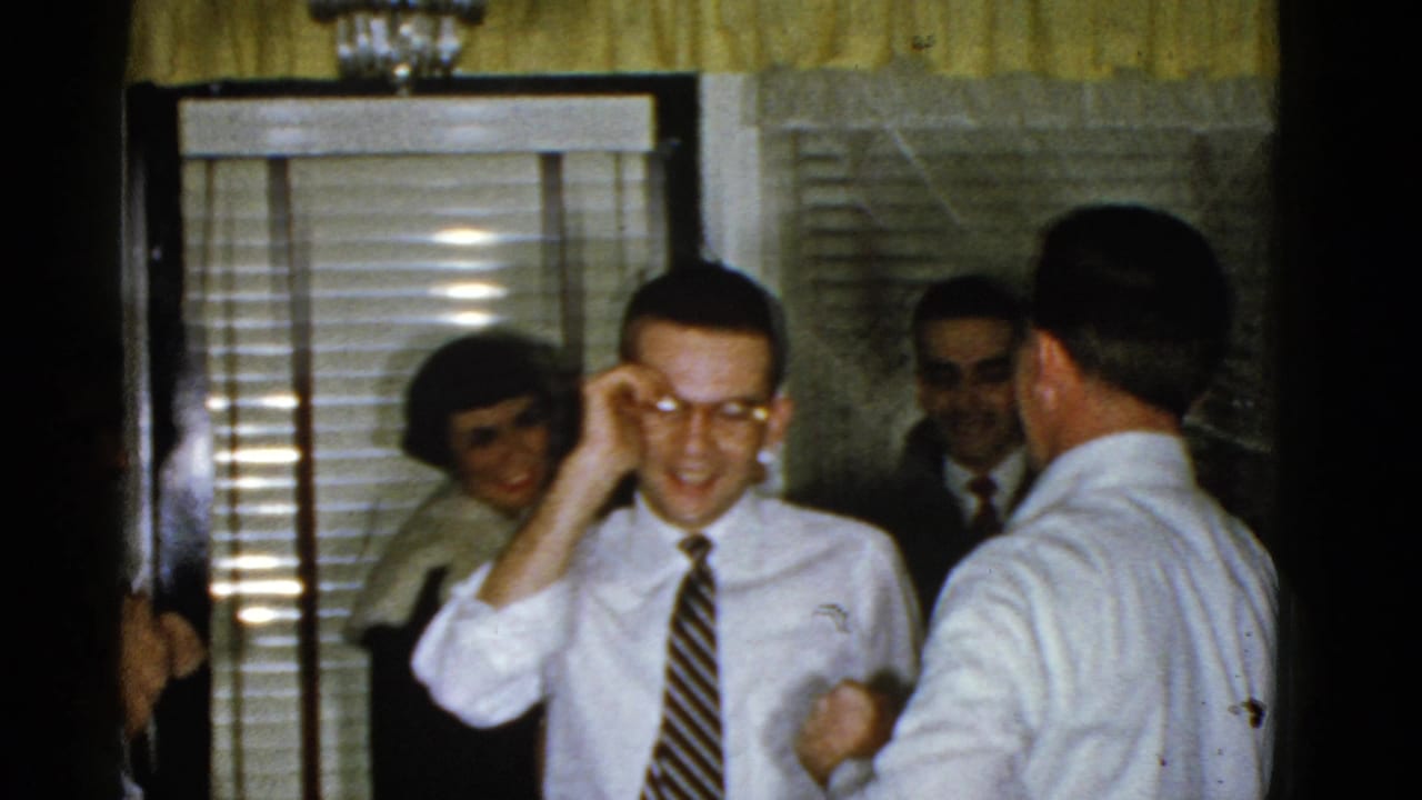 Man in a white shirt adjusts his glasses while other adults stand nearby.