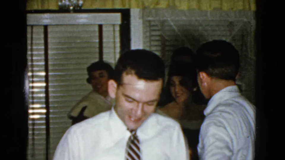 1975 Boston home-movie frame with a smiling man in the foreground and party guests behind him in a private interior.