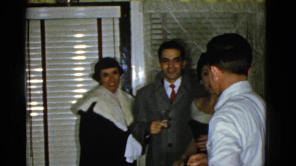 1975 Boston party frame showing adults in suits and dresses, some holding drinks, gathered in a private room.