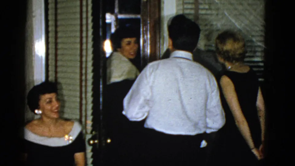 1975 Boston home-movie frame of adults clustered around an interior doorway, including a smiling seated woman at left.