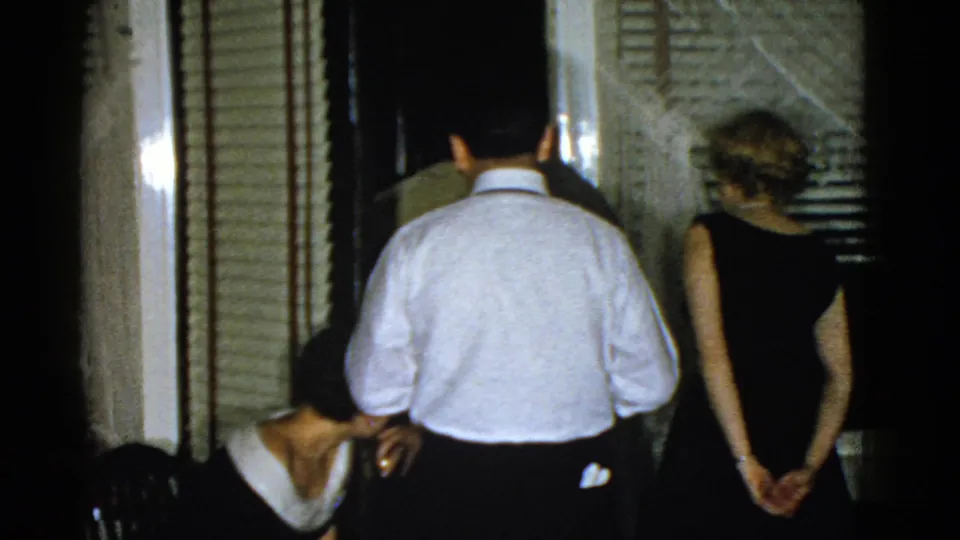1975 Boston home-movie frame of adults departing through an interior doorway while another guest bends down nearby.