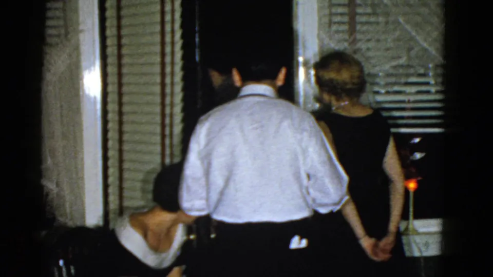 1975 Boston 8mm frame showing adults in formal clothing near a doorway with blinds and curtains during an indoor gathering.