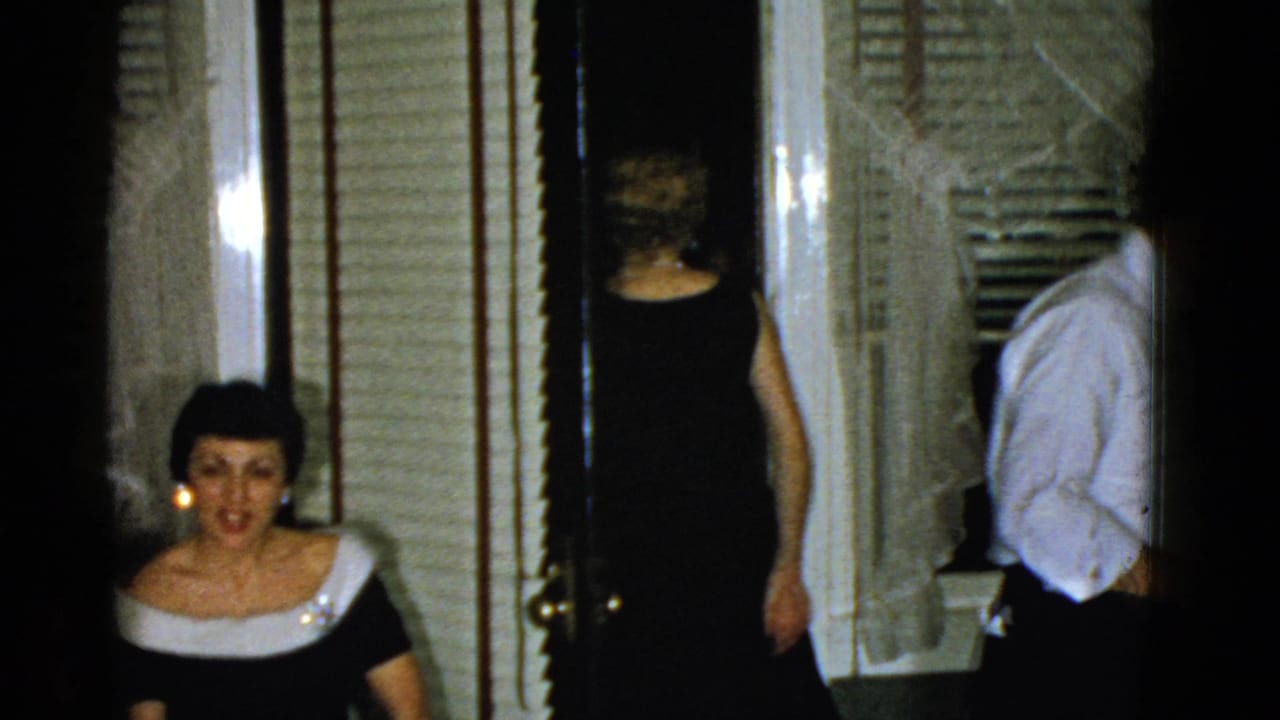1975 Boston home-movie frame with a seated woman in a dark dress with white trim as guests pass through an interior doorway.