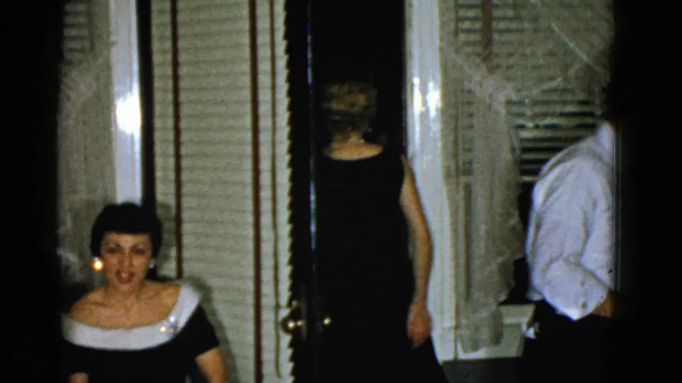 1975 Boston home-movie frame with a seated woman in a dark dress with white trim as guests pass through an interior doorway.