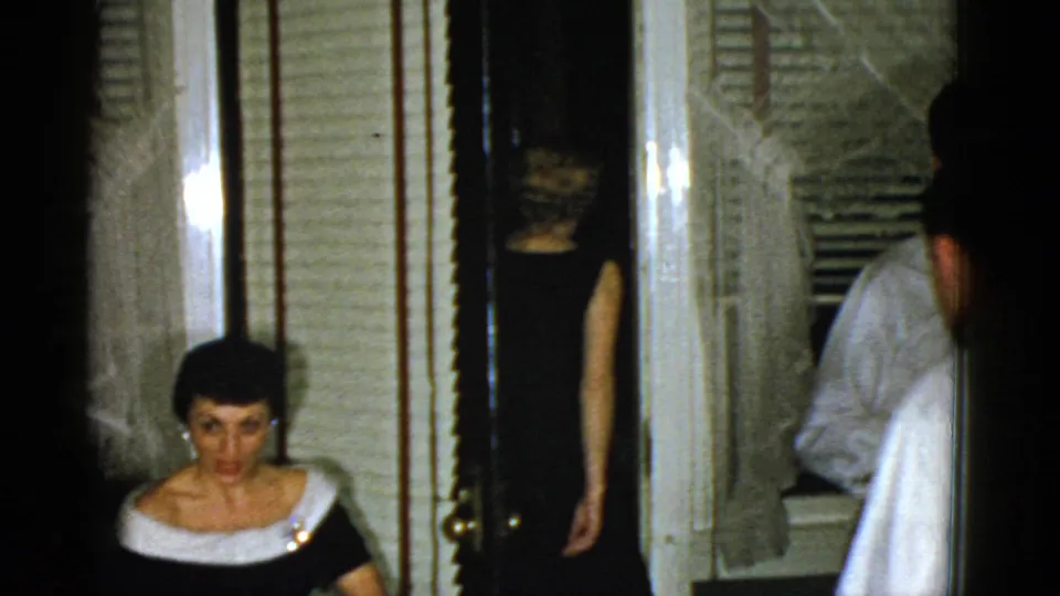1975 Boston 8mm frame showing a seated woman and partially visible guests moving through a dim private interior doorway.