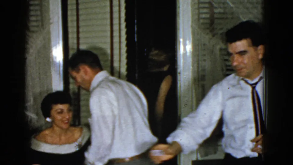 1975 Boston home-movie frame with a man in a white shirt and tie moving past a seated woman near curtains and blinds.