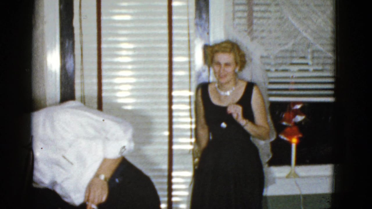 Party guests in a dim room with venetian blinds and a small red lamp.