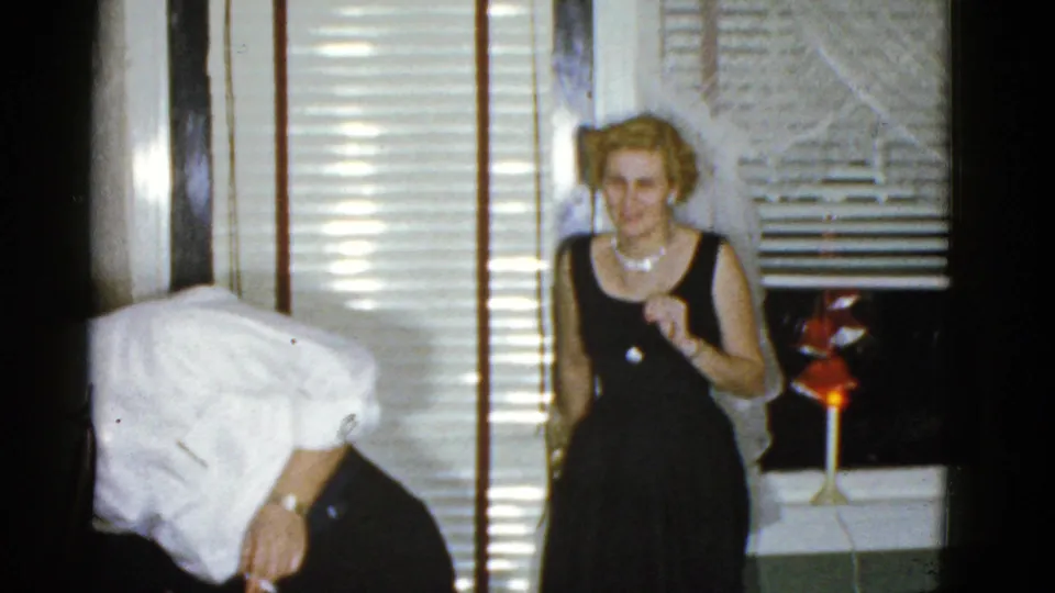 1975 Boston home-movie frame showing adults at an indoor party near venetian blinds, with a woman in a black dress and another guest bending at left.